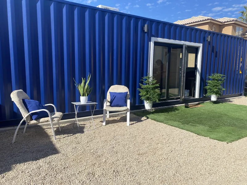 Blue container home with a glass slider opening onto a turf patio with lounge chairs and planters
